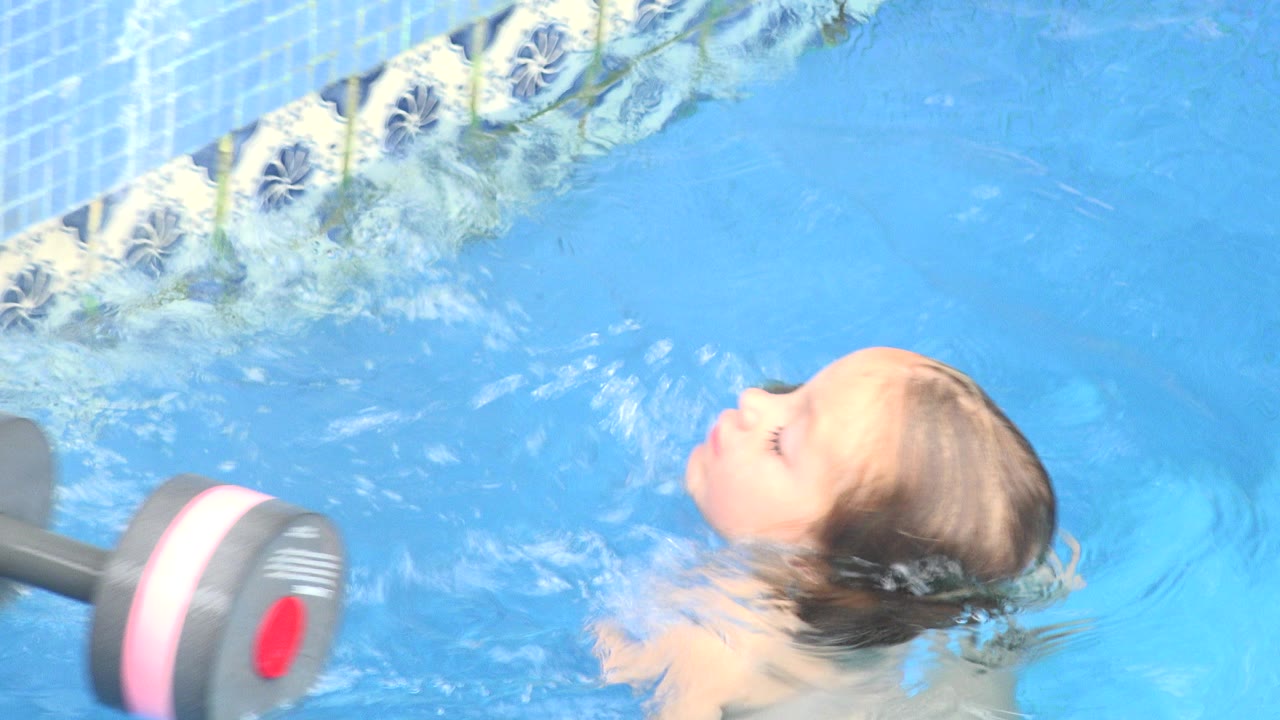 At the start of the Los Cabos pool clip, a young child floats on her back near a gray and pink floating weight, with patterned pool tile along the top edge.