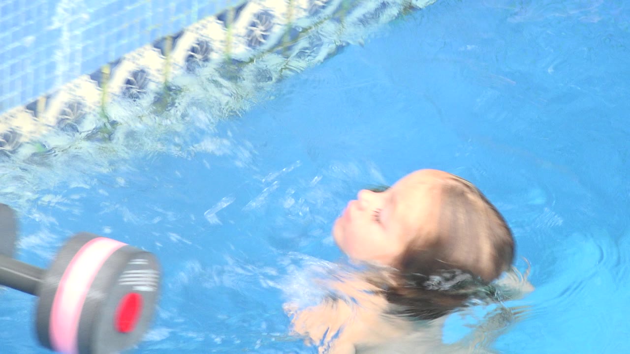 The child continues floating in the Los Cabos pool as small ripples spread around her face and the gray and pink weight sits close to the left foreground.