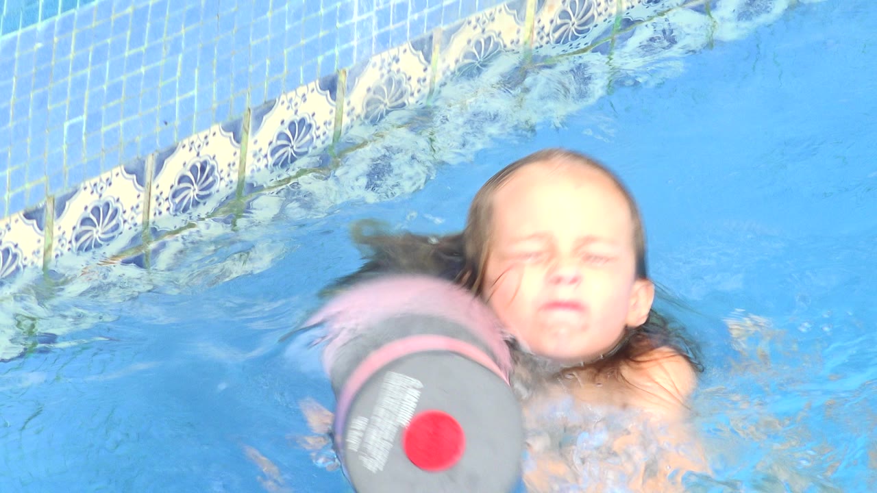 At 0:00 in Los Cabos, Mexico, a young girl keeps her face above bright blue pool water as a gray and pink foam dumbbell float passes close in front of her.