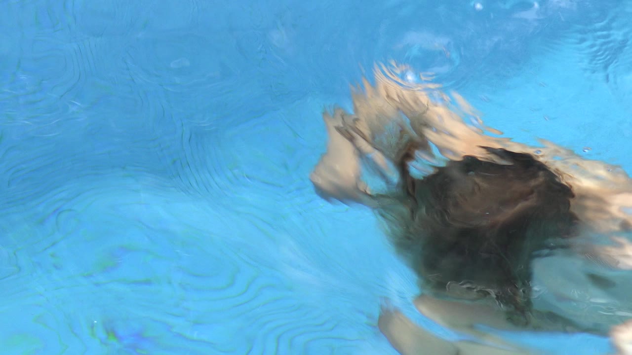 A young child is mostly submerged in blue pool water in Los Cabos, Mexico, with dark hair and a pale arm distorted by ripples near the center of frame.