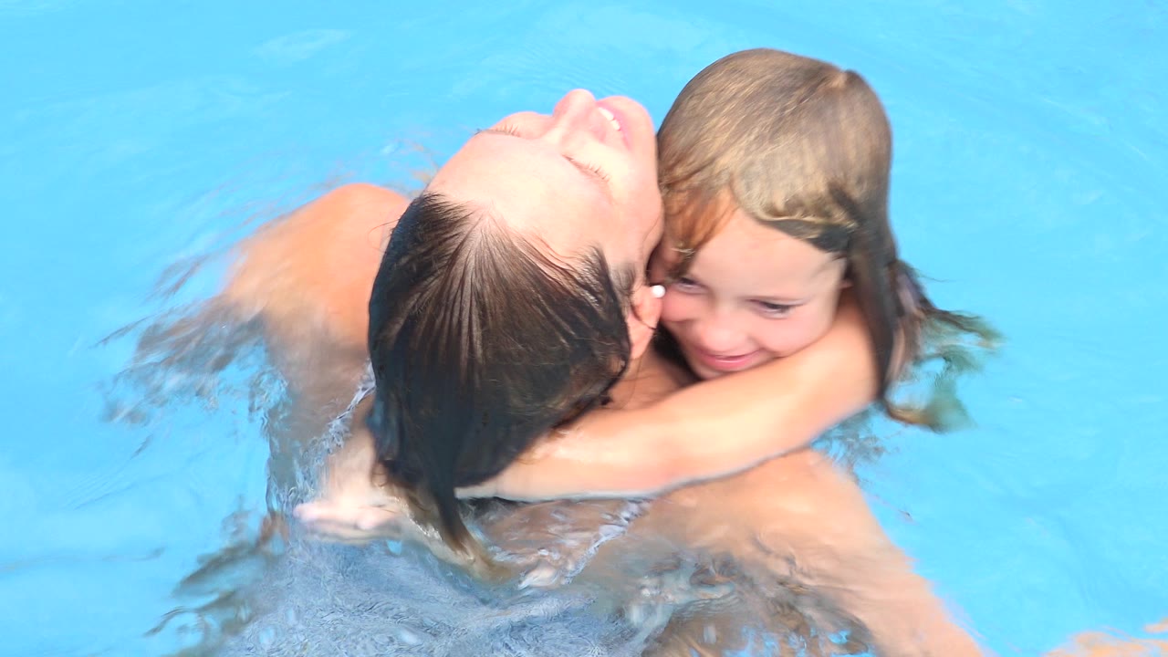 In Los Cabos, Mexico, the young girl smiles while hugging the woman from the front in clear blue pool water.