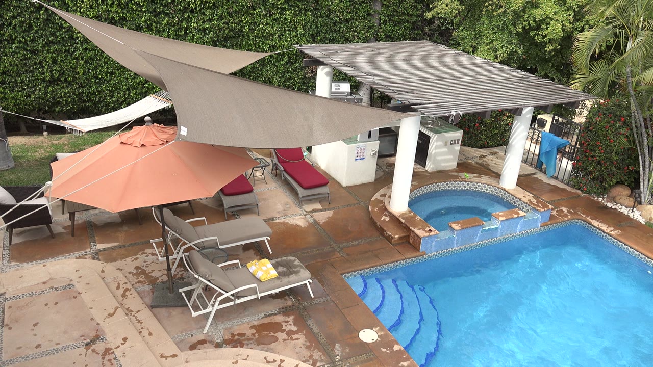 Los Cabos pool patio frame with the blue pool at lower right, wet tile marks, recliners, a coral umbrella, shade sails, white columns, and no visible people.