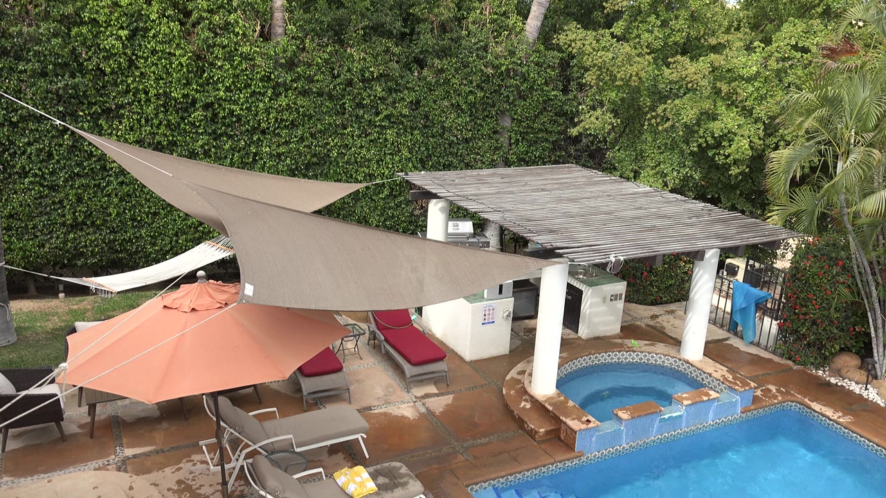Elevated Los Cabos view of a blue pool edge, round spa, orange umbrella, beige shade sail, red-cushioned loungers, palms, and dense hedges, with no visible people.