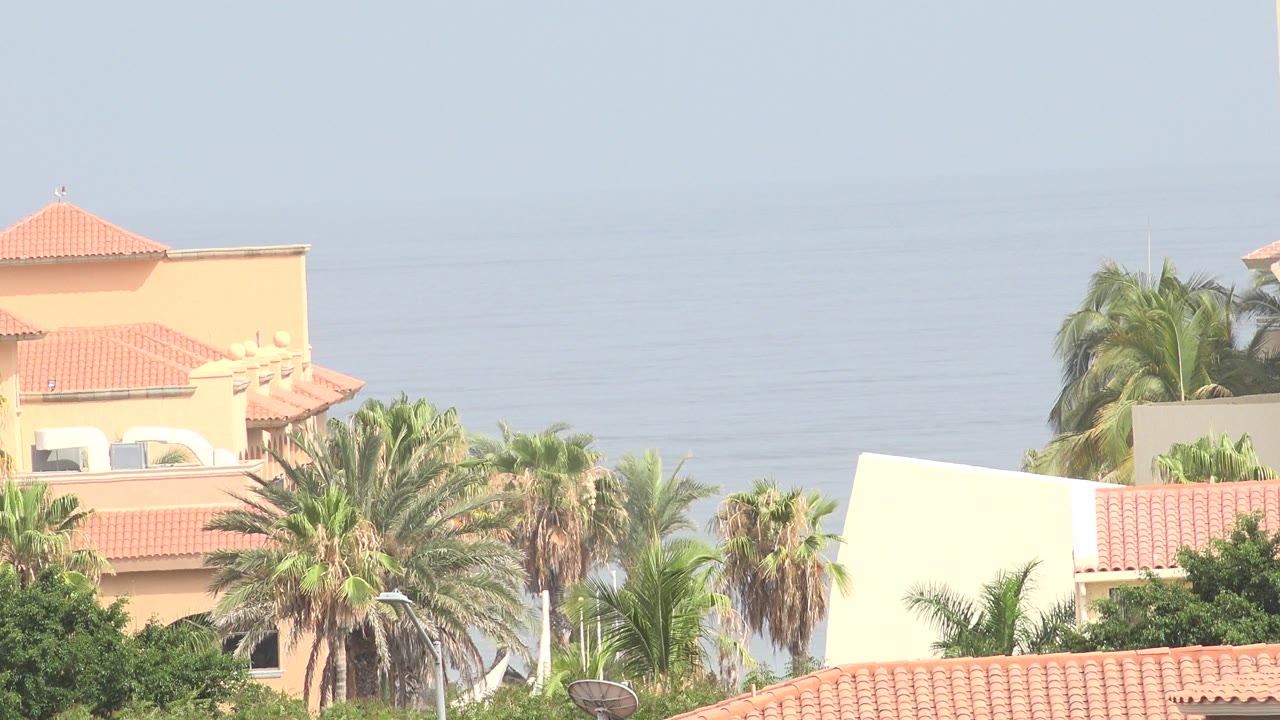 Opening frame of Los Cabos, Mexico homes with red tile roofs, palm foliage, pale ocean haze, and no visible people.