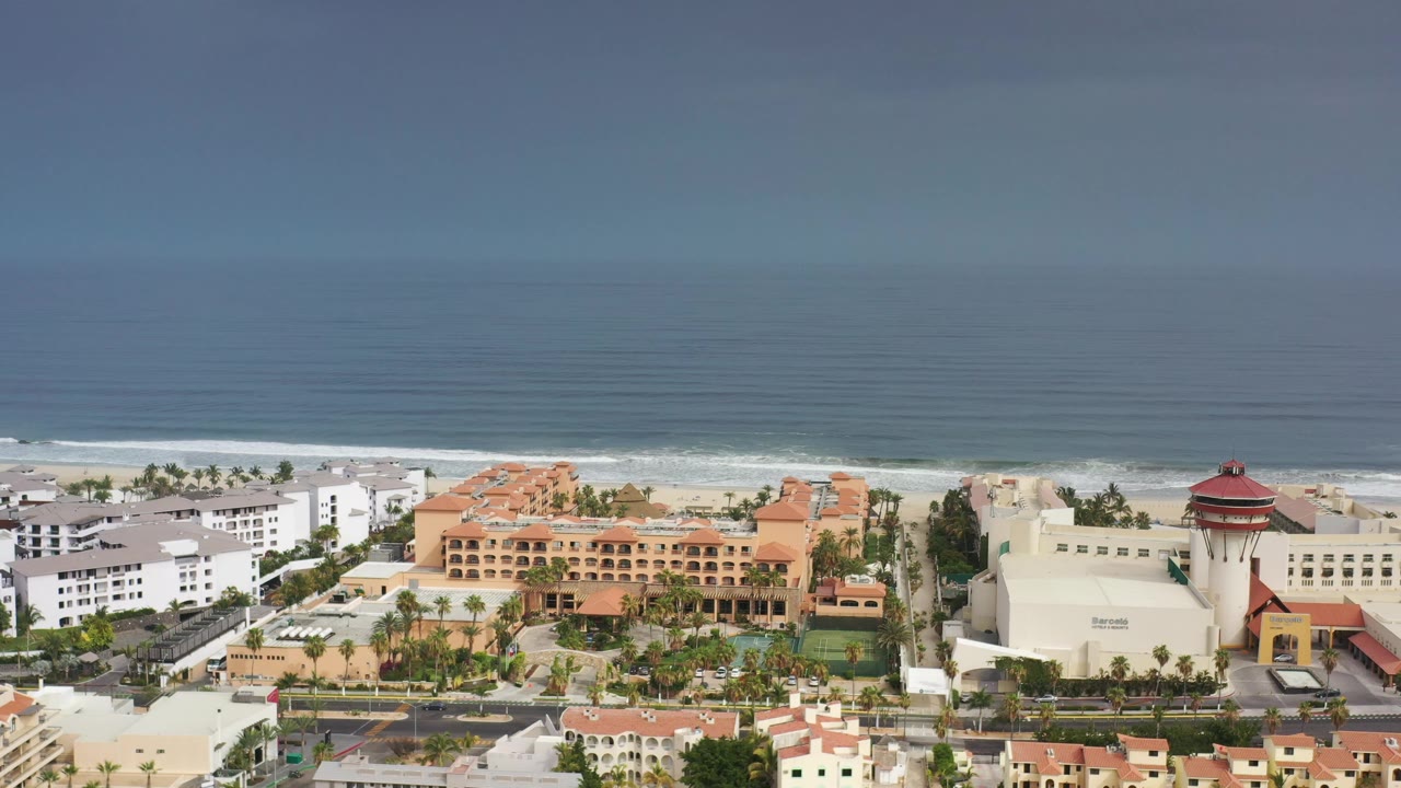 Los Cabos aerial frame showing a white building cluster, orange-roofed beachfront structures, a road with cars, the surf line, and no visible people.