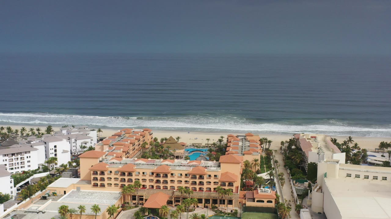 Aerial view of beachfront buildings, sand, surf, and ocean horizon.