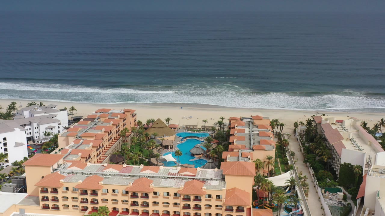 Opening aerial frame over a Los Cabos resort with terracotta roofs, blue pools, palm landscaping, a pale beach, rolling surf, and tiny distant people on the sand.