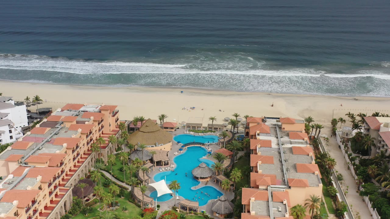 Aerial view of beachfront resort buildings, pools, sandy beach, and ocean surf.