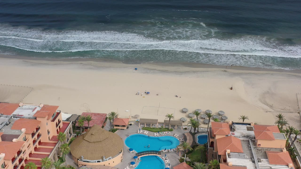 Aerial view of surf, sand, beachfront buildings, pools, and palms.