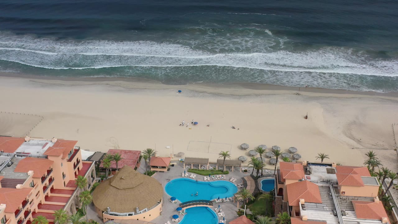 Overhead Los Cabos coastline frame with breaking waves, pale open sand, a cluster of beachfront roofs, bright pool water, palms, and no visible people.