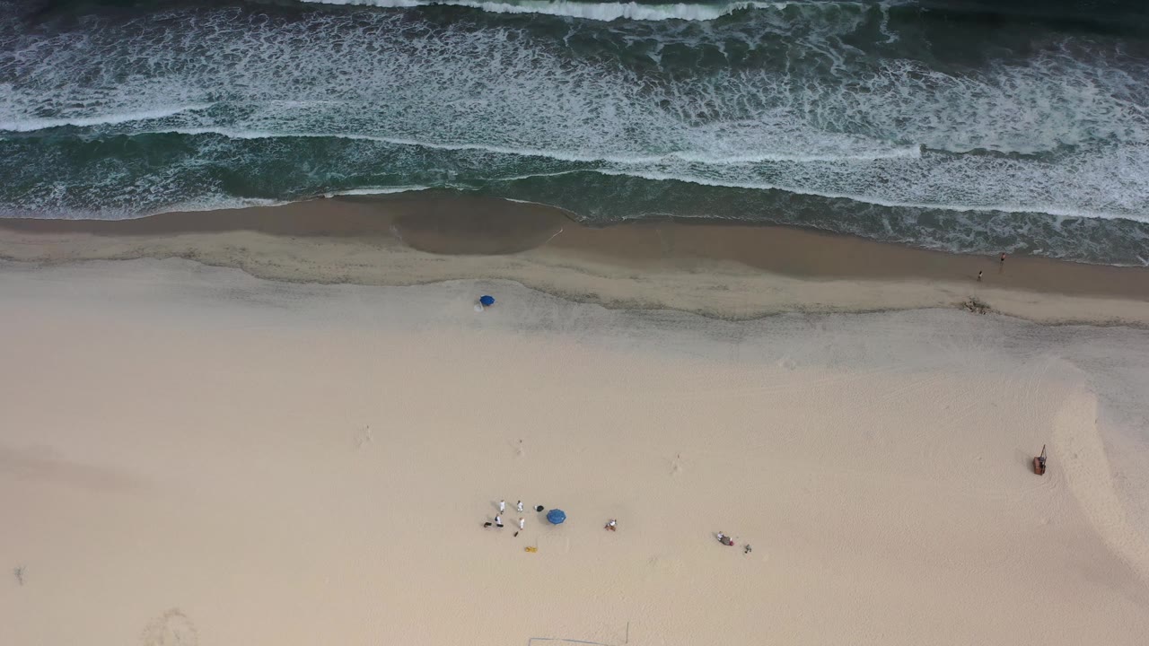 Aerial Los Cabos beach frame with whitewater near the top, broad tan sand below, and small blue beach items visible, with no visible people.