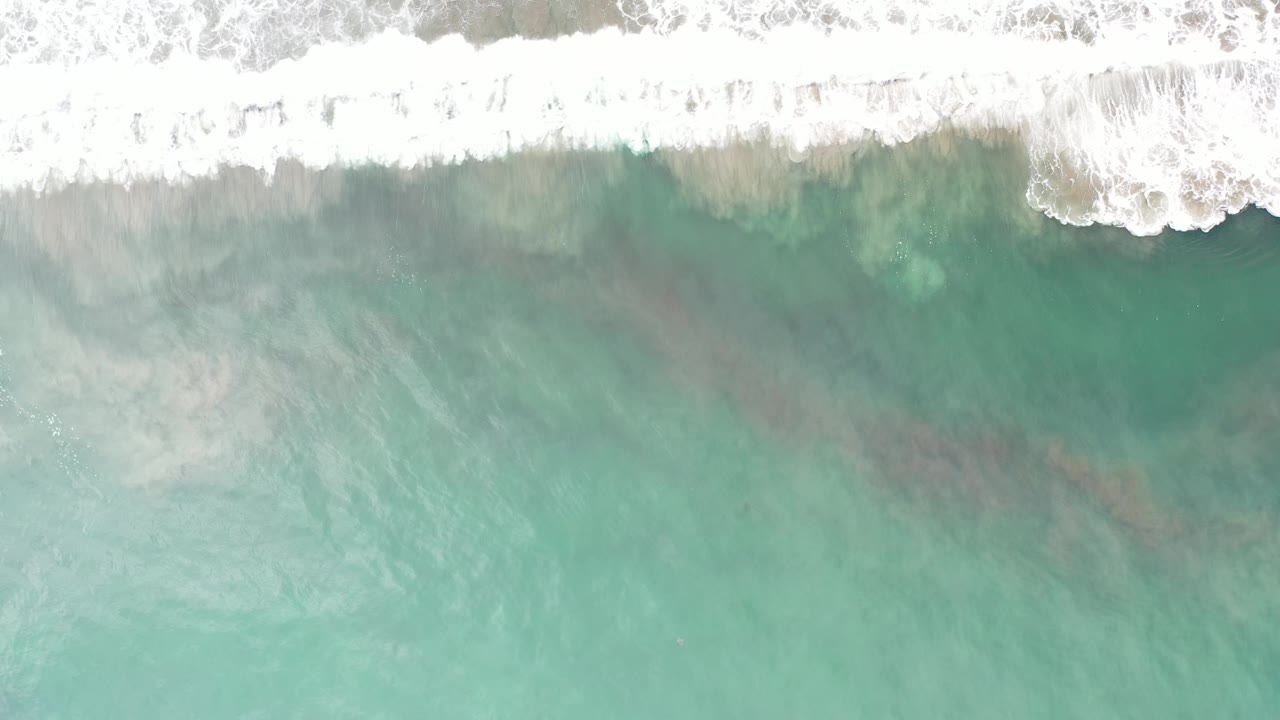 Aerial frame of Los Cabos green sea with white breaking foam along the upper border and faint darker patches under the water, with no visible people.