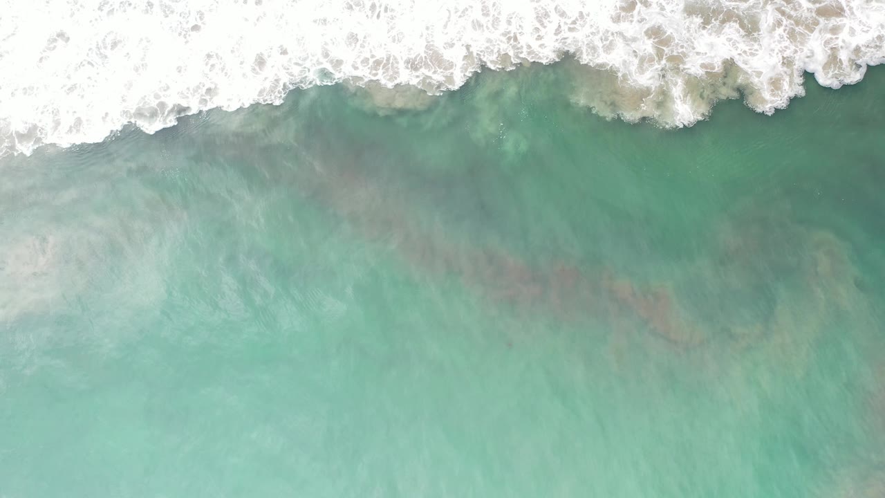 White foamy wash spreads across the upper edge while pale turquoise water and sand tones fill the lower frame in Los Cabos, Mexico, with no visible people.