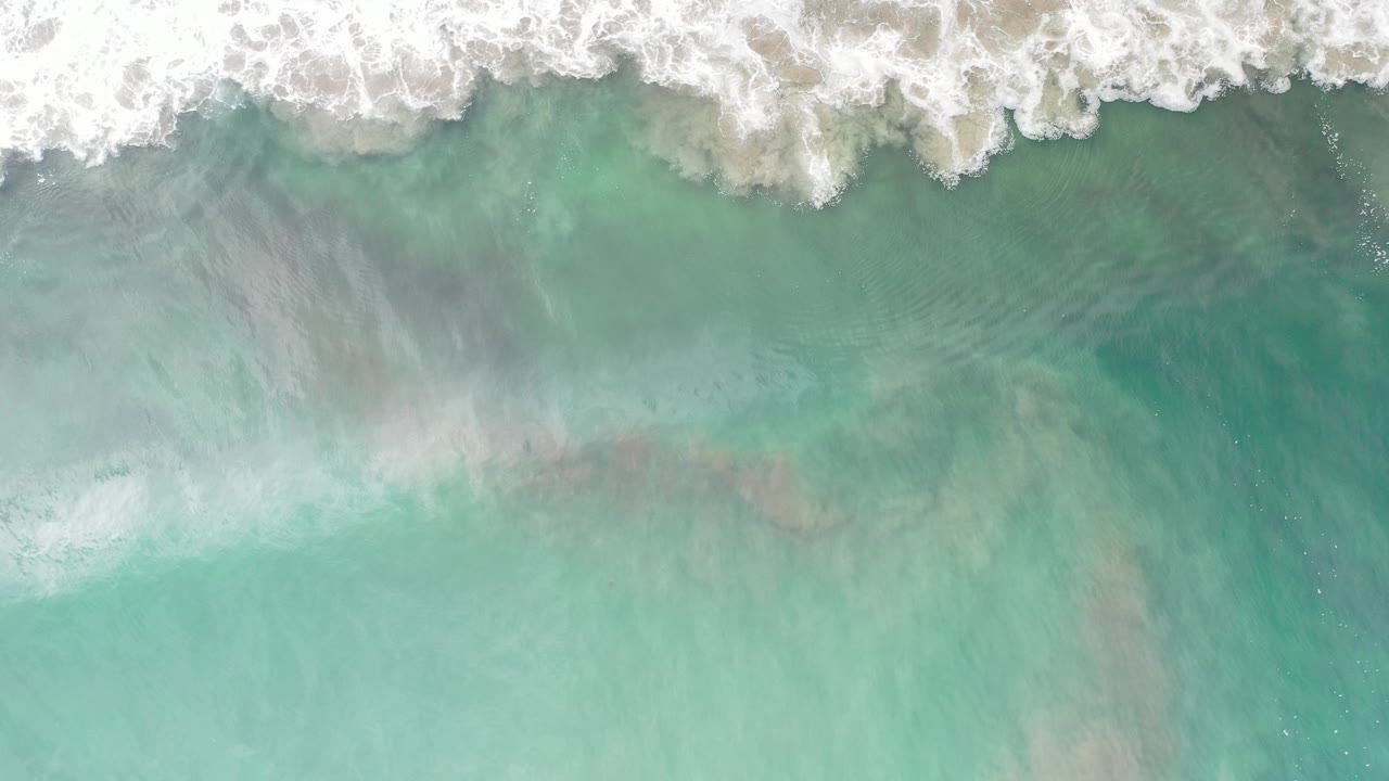 Overhead view of turquoise ocean water and white surf in Los Cabos, Mexico, with no visible people at the start of the clip.
