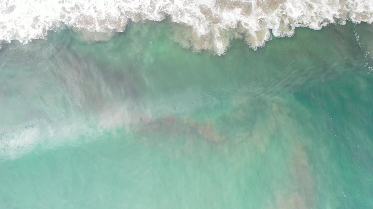 Foamy surf forms a bright band above clear blue-green water in Los Cabos, Mexico, with no visible people.