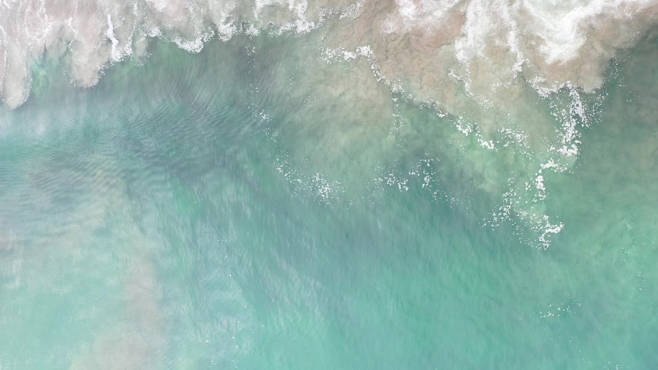 Overhead view of pale green seawater and white surf foam along the upper edge in Los Cabos, Mexico, with no visible people.