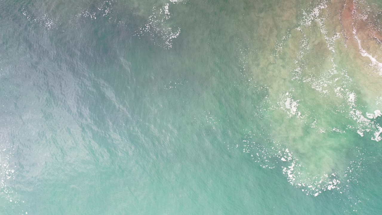 Top-down Los Cabos ocean view with turquoise water, pale submerged coastal texture near the upper right, scattered foam, and no visible people.