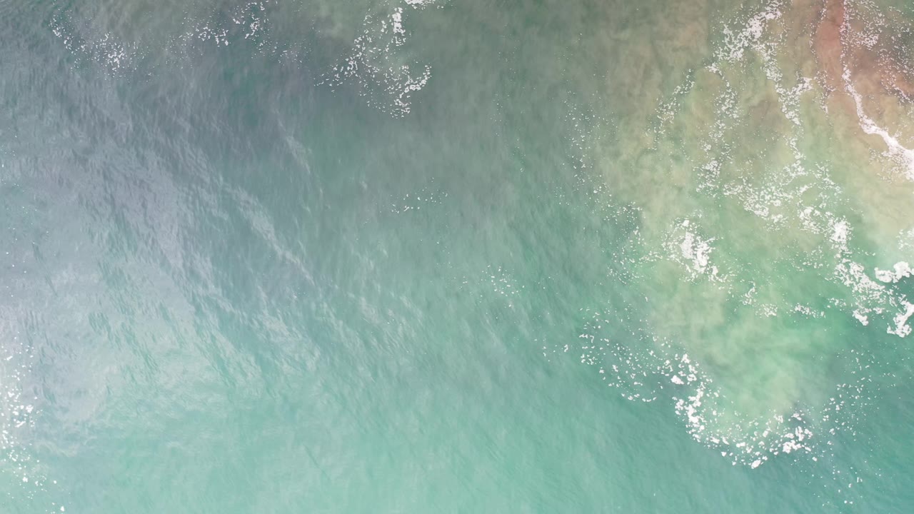 Los Cabos aerial water frame showing green-blue ocean, a pale rocky or sandy patch near the right side, light foam clusters, and no visible people.