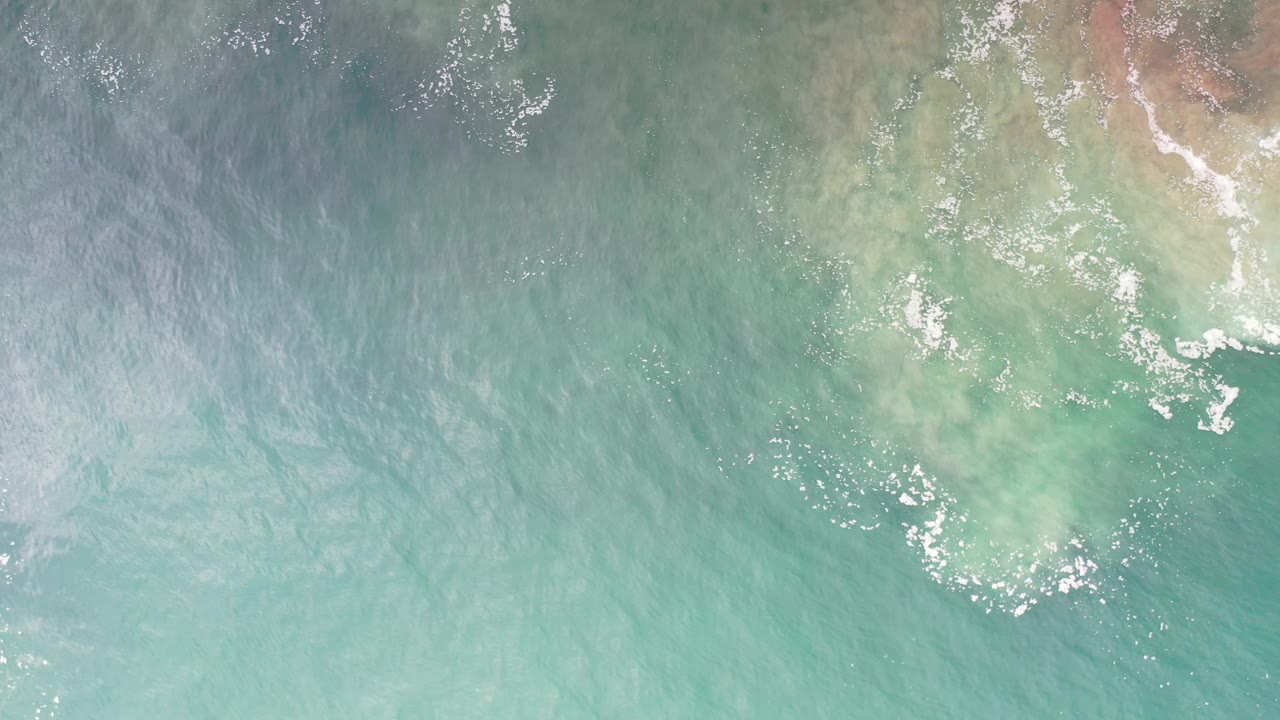 Overhead sea water with a pale coastal patch and foam near the upper right.