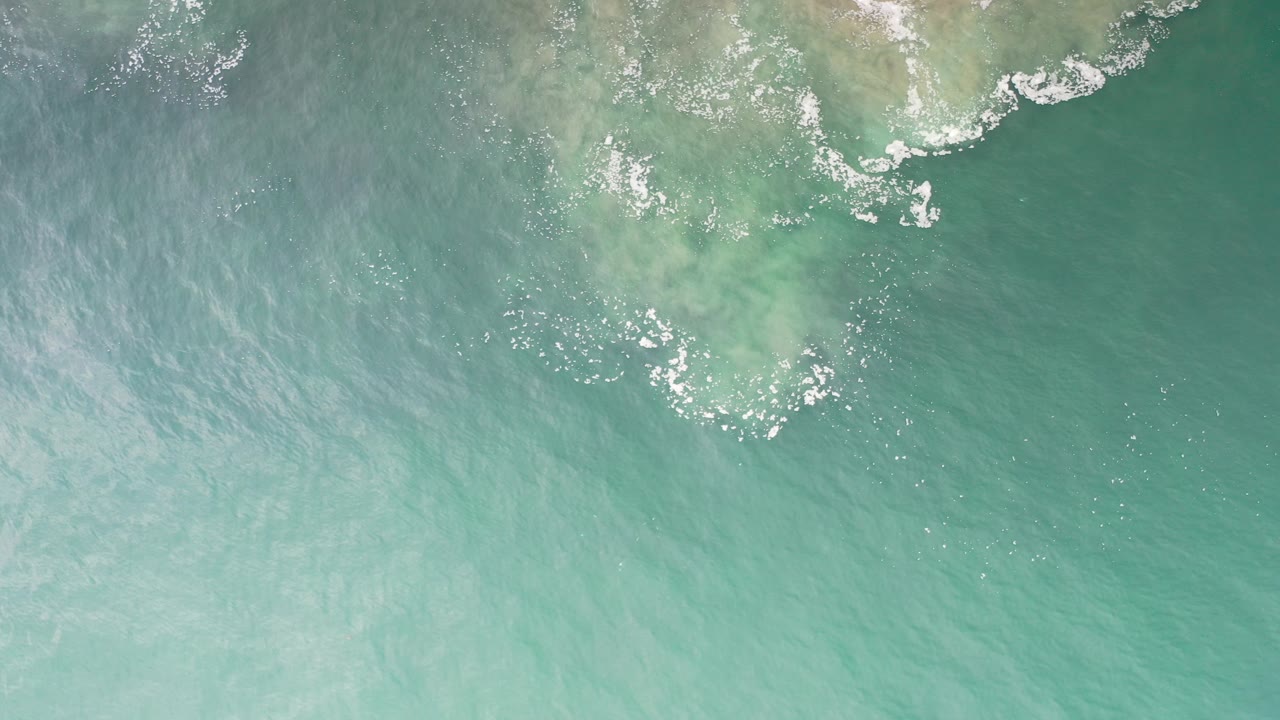 Overhead view of green sea water in Los Cabos, Mexico, with pale sand visible beneath foam near the top edge and no visible people.