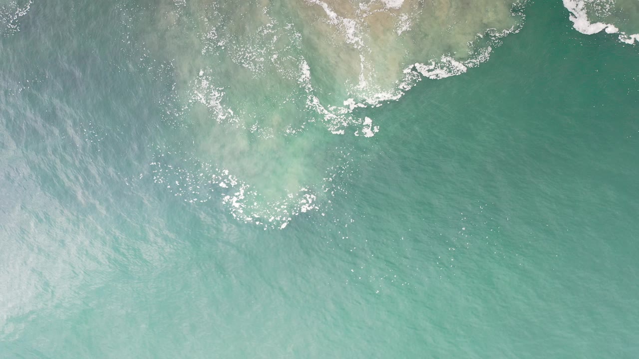 Overhead green ocean water with white foam near pale shoreline patches.