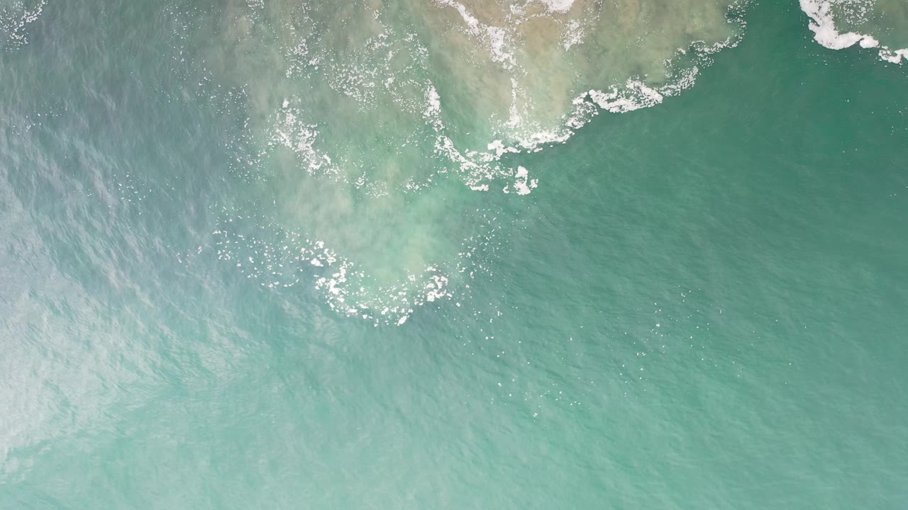 Aerial frame over turquoise-green water in Los Cabos, Mexico, where white foam breaks around pale shallow patches near the upper edge and no visible people appear.