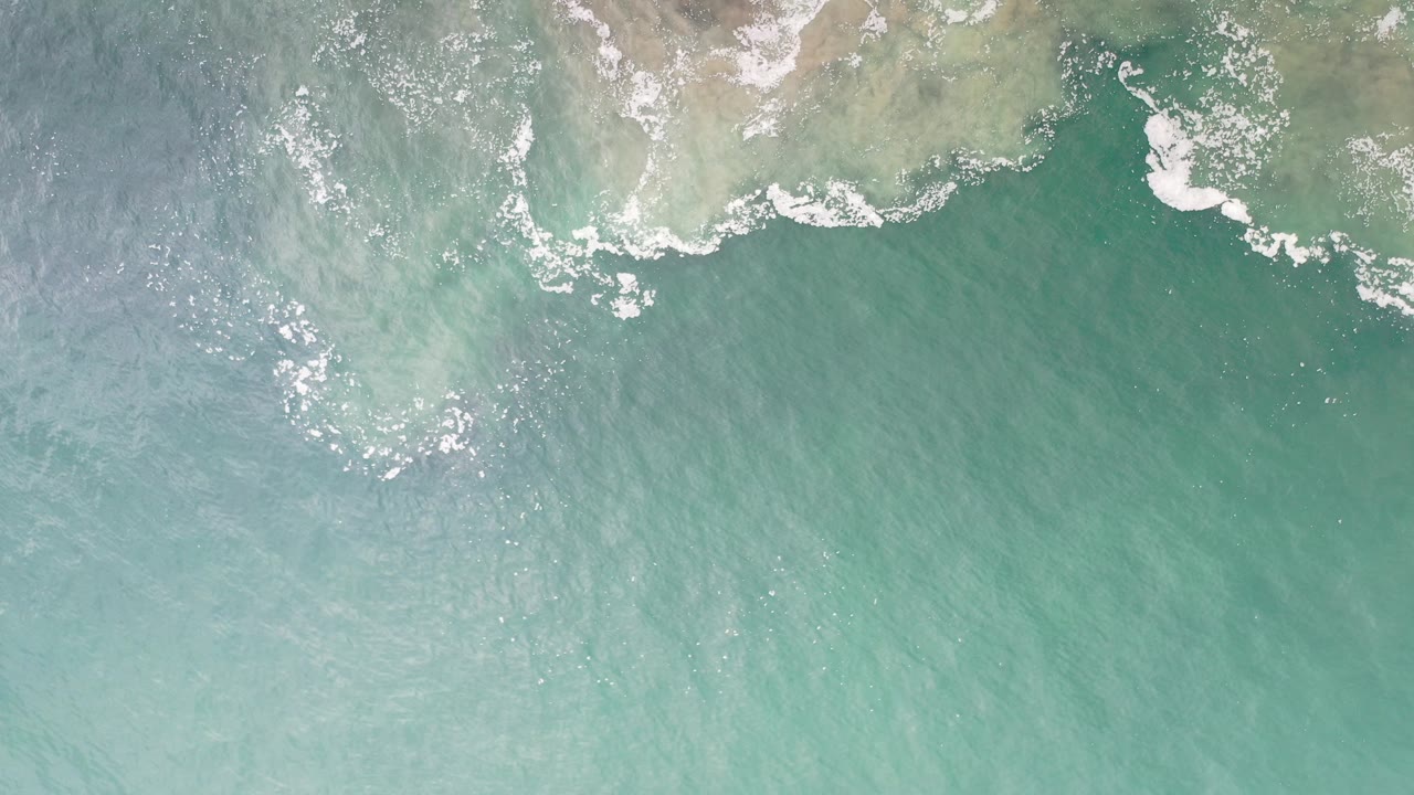 Overhead view of turquoise-green water meeting a pale Los Cabos shoreline with white foam at the edge and no visible people.