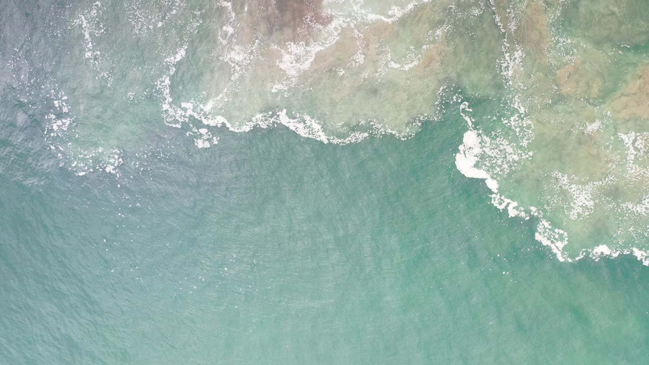 Overhead view in Los Cabos, Mexico of turquoise water and pale rocky shallows with white surf along the upper edge, with no visible people.