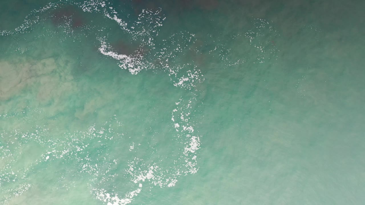 Overhead view of turquoise seawater in Los Cabos, Mexico, with white foam curling across the upper frame and no visible people.