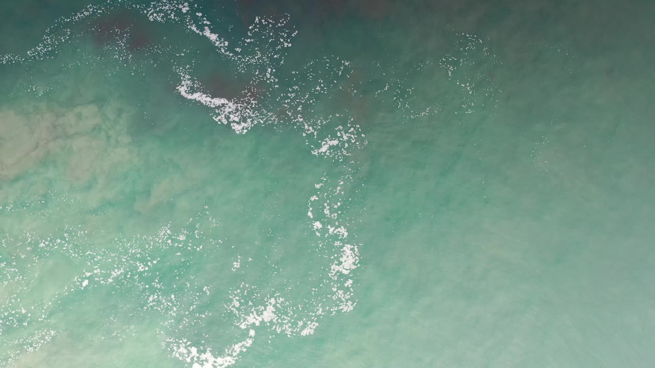Top-down Los Cabos ocean frame with pale turquoise water, a curved white foam line near the top, and no visible people.
