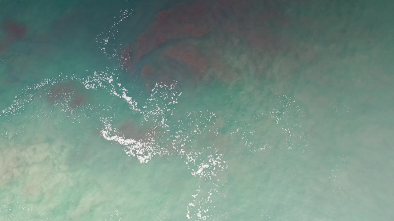 Overhead view of turquoise ocean water in Los Cabos, Mexico, with white foam near the left side, darker blue water above, and no visible people.