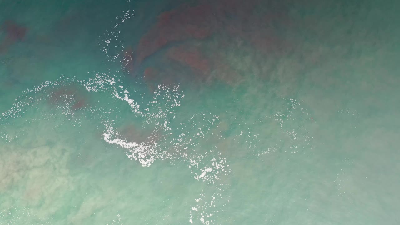 Top-down blue-green sea surface in Los Cabos, Mexico, with scattered white foam, reddish-brown underwater patches, and no visible people.