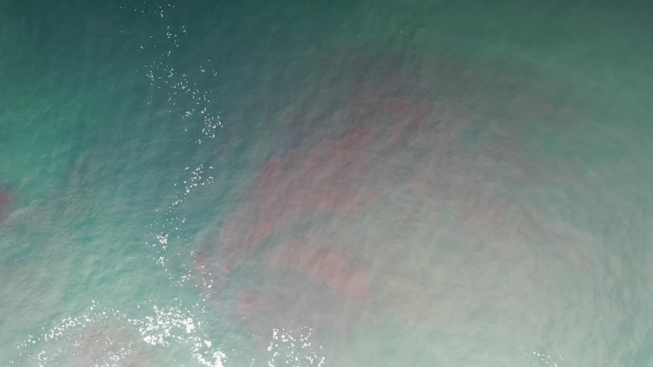 Overhead view of blue green ocean water in Los Cabos, Mexico, with a broad pale foamy area and faint pink reflection near the lower frame, no visible people.