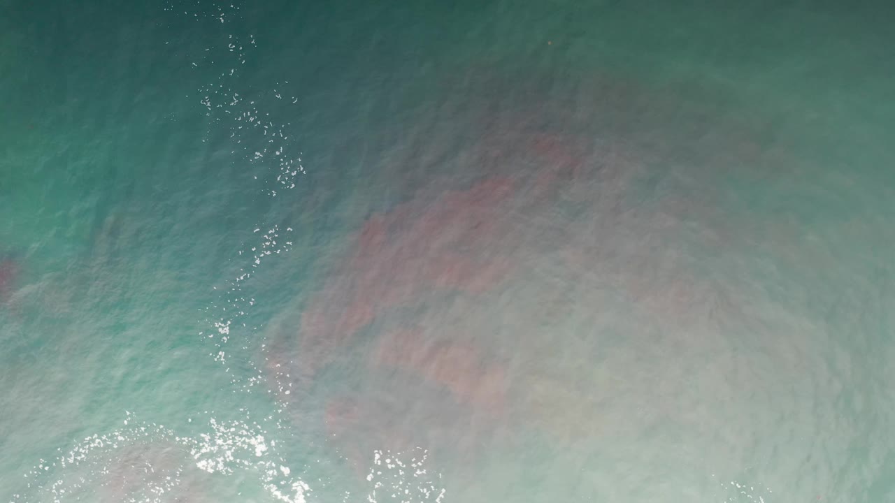 Overhead Los Cabos ocean surface with turquoise water, cloudy white foam, and muted red pink streaks spreading across the center, no visible people.