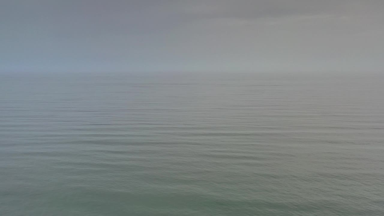 Wide Los Cabos ocean view at the start of the clip, with muted gray-blue sky, a soft horizon, rippled green-gray water, and no visible people.