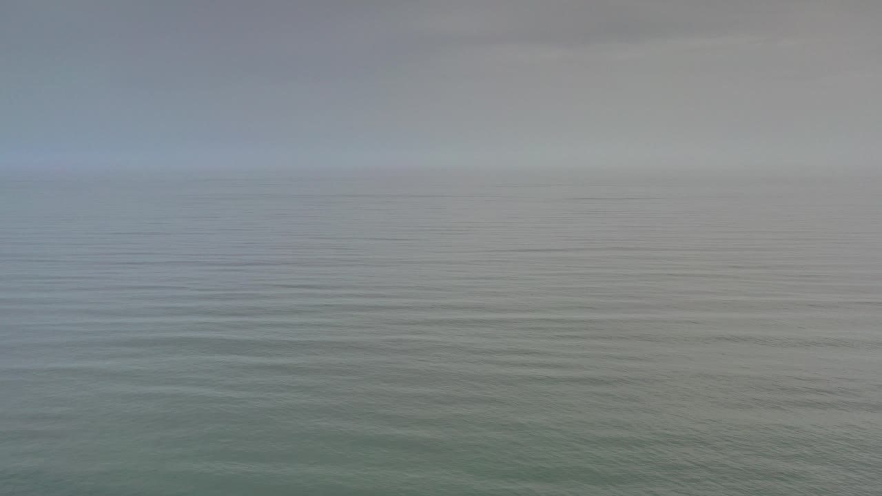 Opening view of the Los Cabos sea under hazy cloud cover, with muted gray-green water and no visible people.
