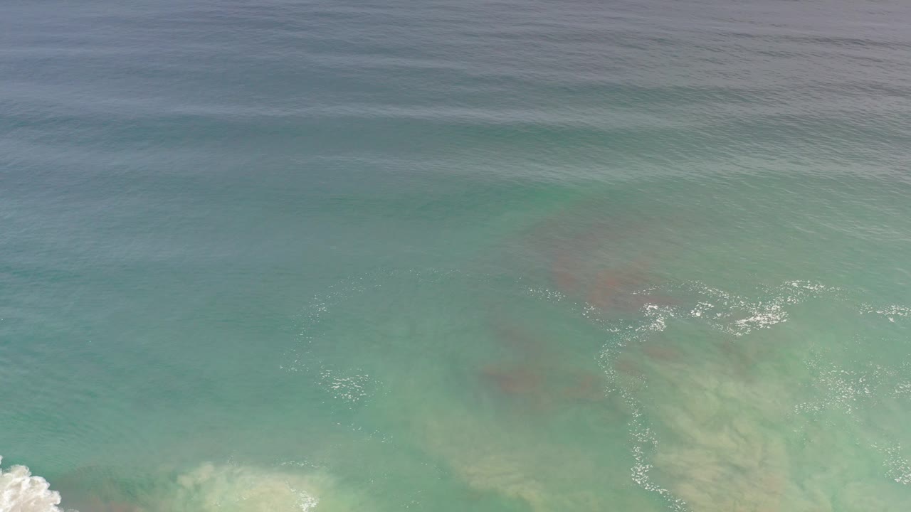 Aerial view of clear green-blue ocean in Los Cabos, Mexico, with pale sand visible under the water, small foam traces near the lower left, and no visible people.