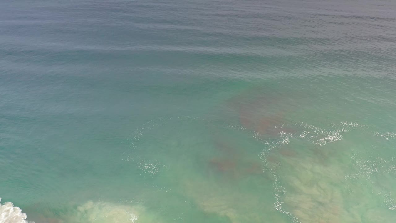 Aerial frame over Los Cabos water, showing translucent green surf, darker submerged areas, white ripples near the lower right, and no visible people.