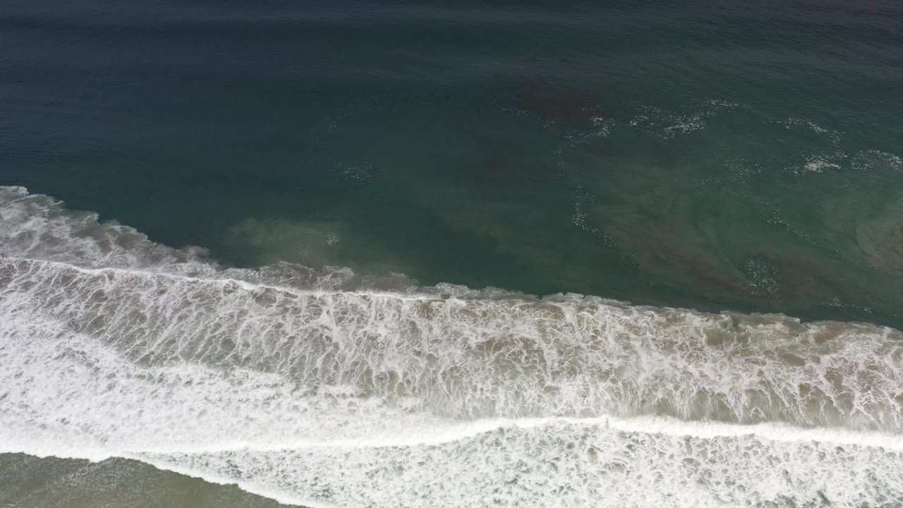 Overhead view of white surf rolling across dark teal water toward a sandy Los Cabos beach, with no visible people.
