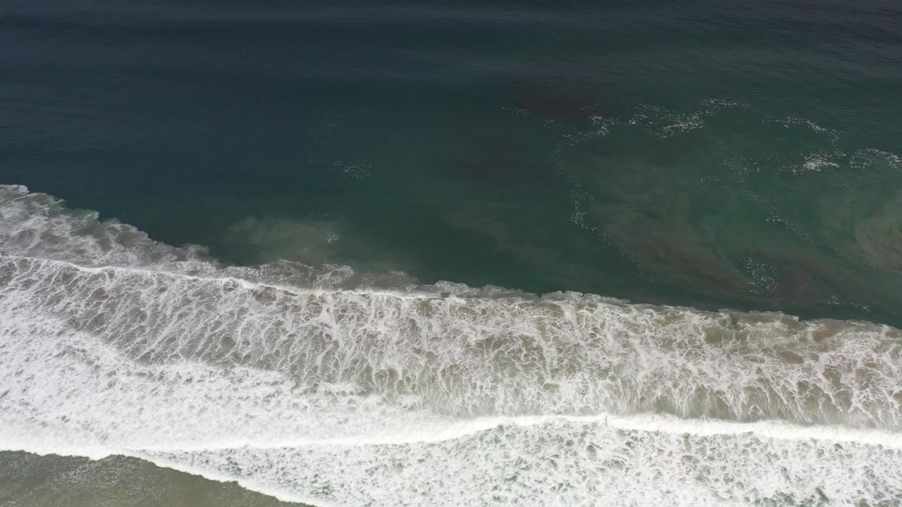Aerial frame of broad foam lines stretching across green-blue water above the sand in Los Cabos, with no visible people.