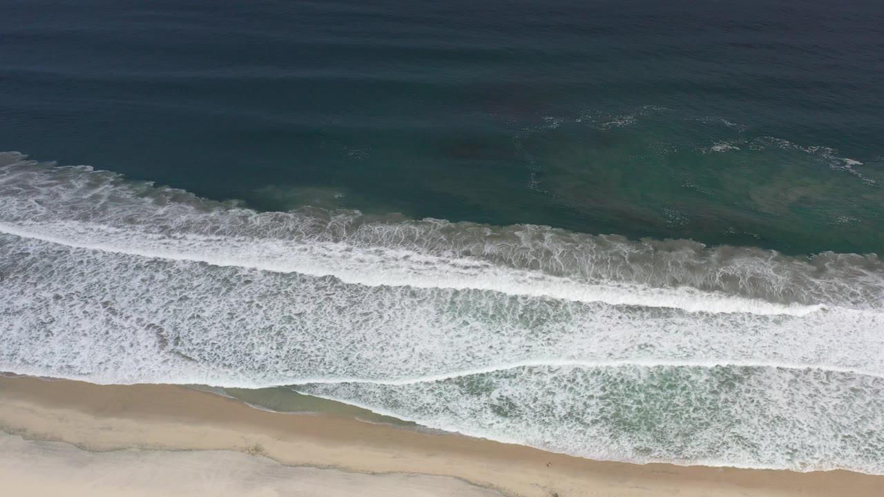 Overhead Los Cabos coastline view with dark blue ocean, green shallows, white surf, pale sand, and no visible people at the start of the clip.