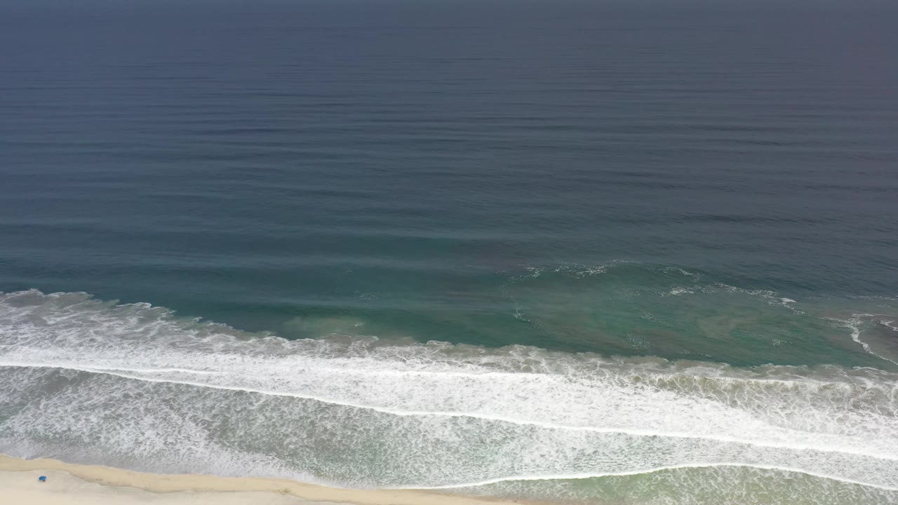 The Los Cabos shoreline remains seen from above as a broad white foam band rolls across green water with a small strip of sand and no visible people.