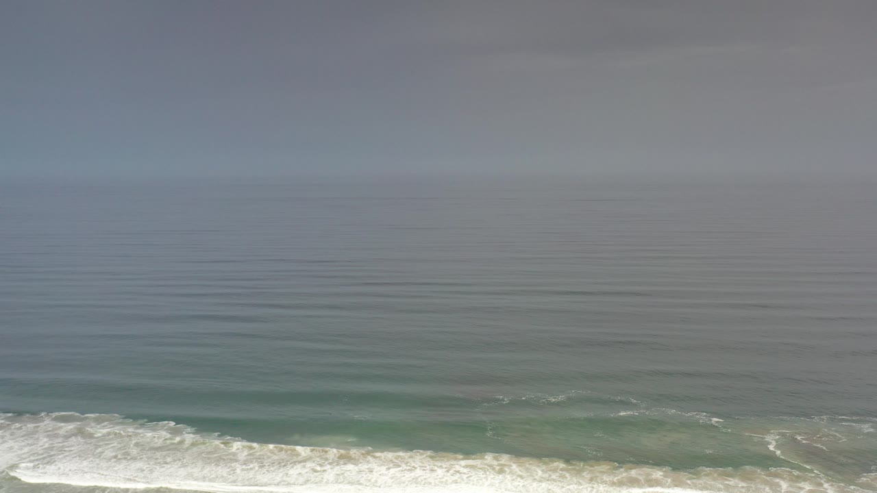 Opening aerial frame over the blue-green ocean in Los Cabos, Mexico, with white surf along the lower edge, a hazy horizon, and no visible people.
