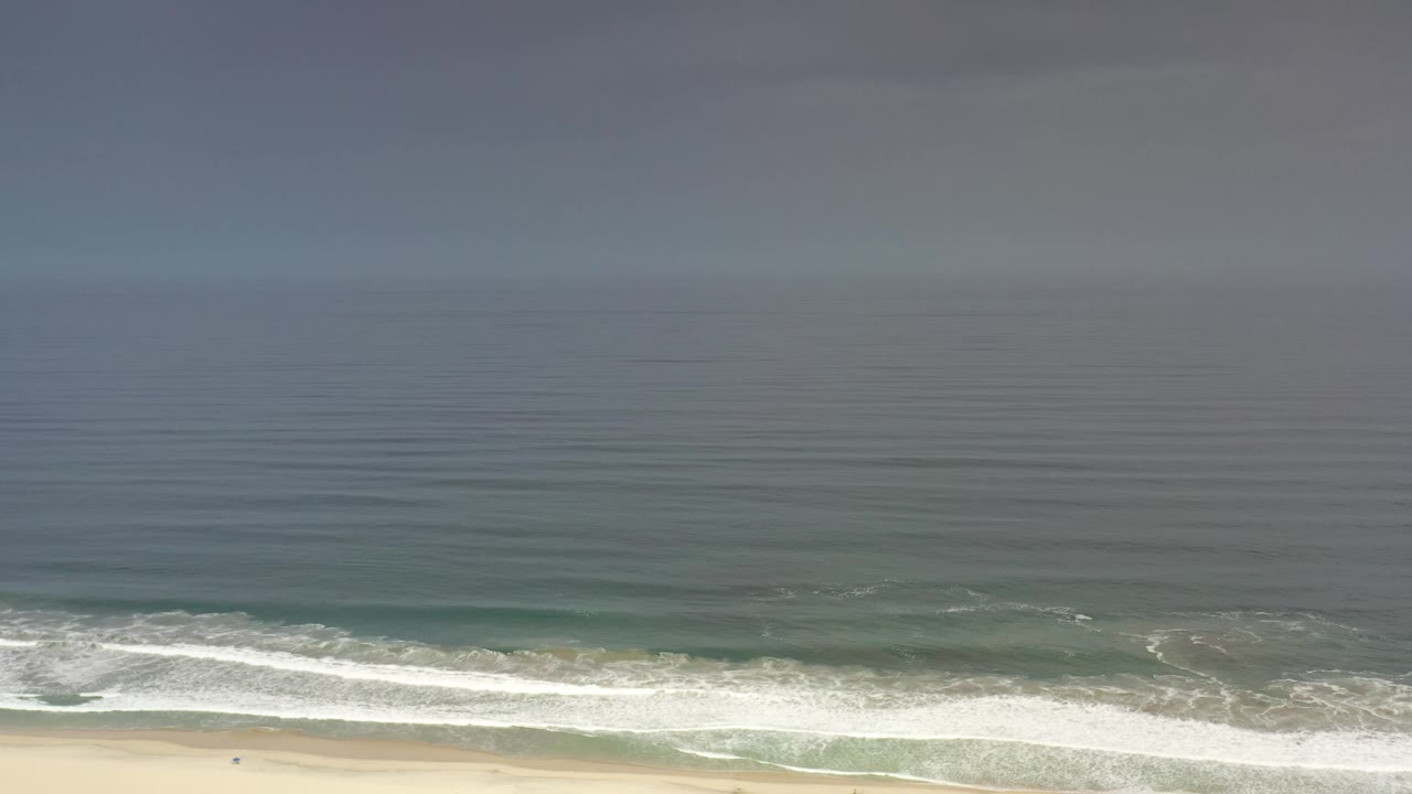 The Los Cabos shoreline remains distant from the aerial camera as a brighter foam line spreads across the lower part of the blue ocean, with no visible people.