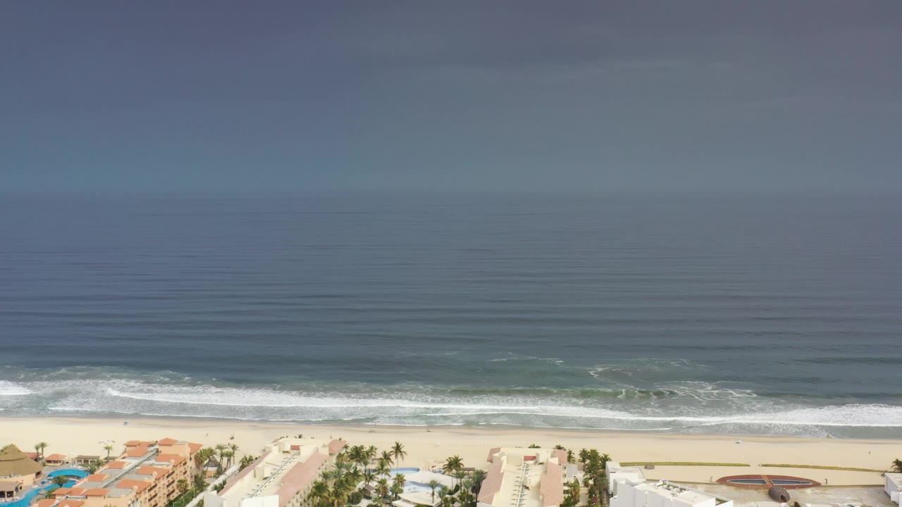 At 0:01, the Los Cabos shoreline remains wide in frame as gray-blue water forms horizontal bands and surf rolls toward the beach, with no visible people.