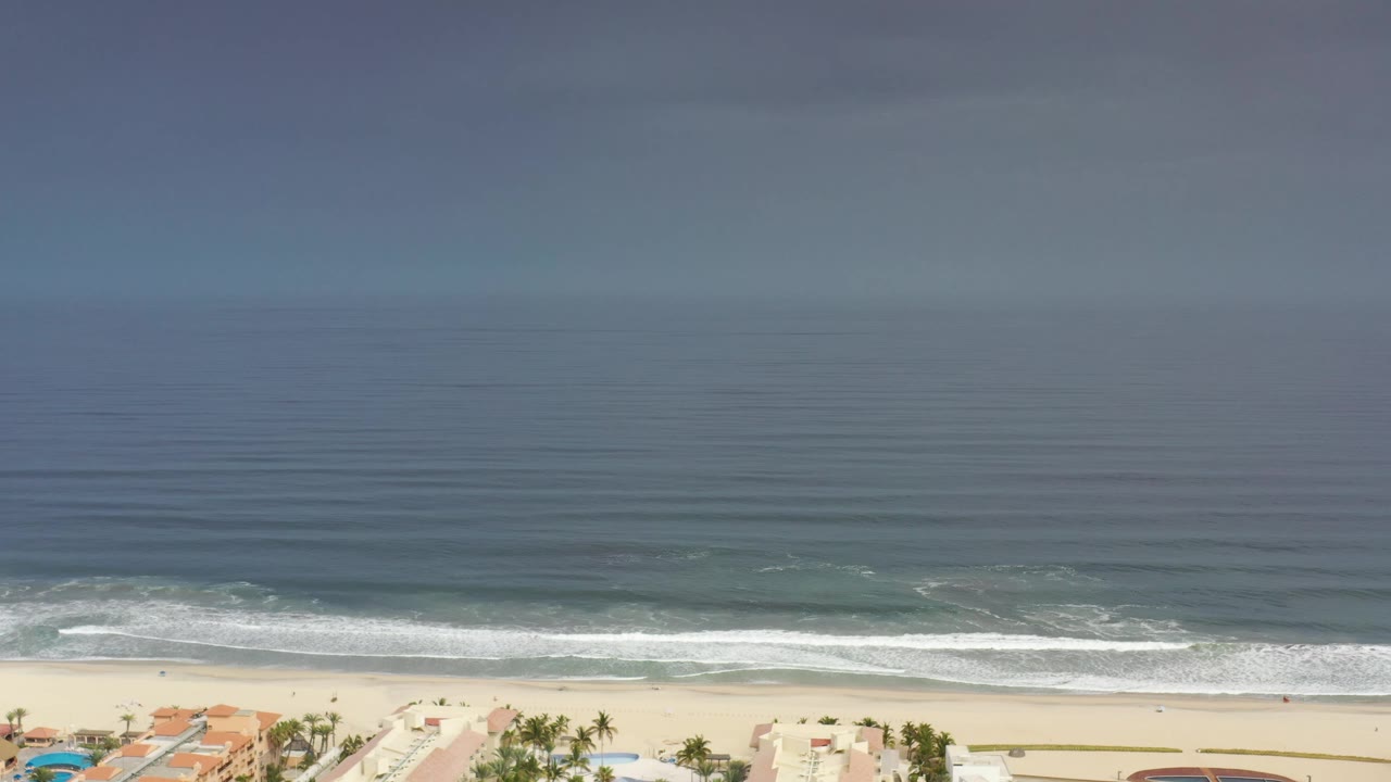 Wide Los Cabos aerial frame with open water across the center, low white waves near shore, pale sand, and small rooftops along the lower edge, no visible people.