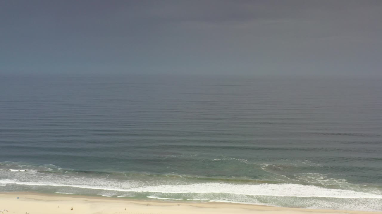 Opening frame of Los Cabos shoreline with blue-green water, white surf bands, sandy beach at the bottom edge, and tiny distant people visible near the waterline.