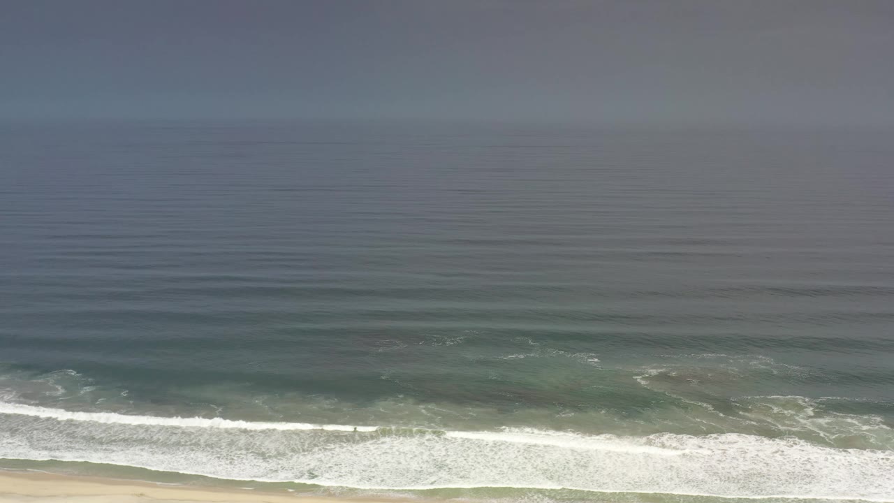 Los Cabos shoreline frame showing layered blue green water, a bright surf line, sand at the lower edge, and no visible people.