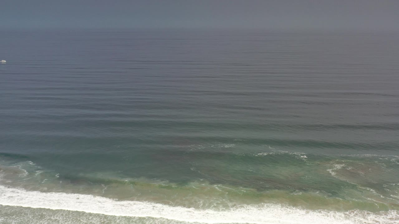 Elevated Los Cabos ocean view with blue-green water, white surf entering along the lower edge, a hazy horizon, a tiny distant boat, and no visible people.