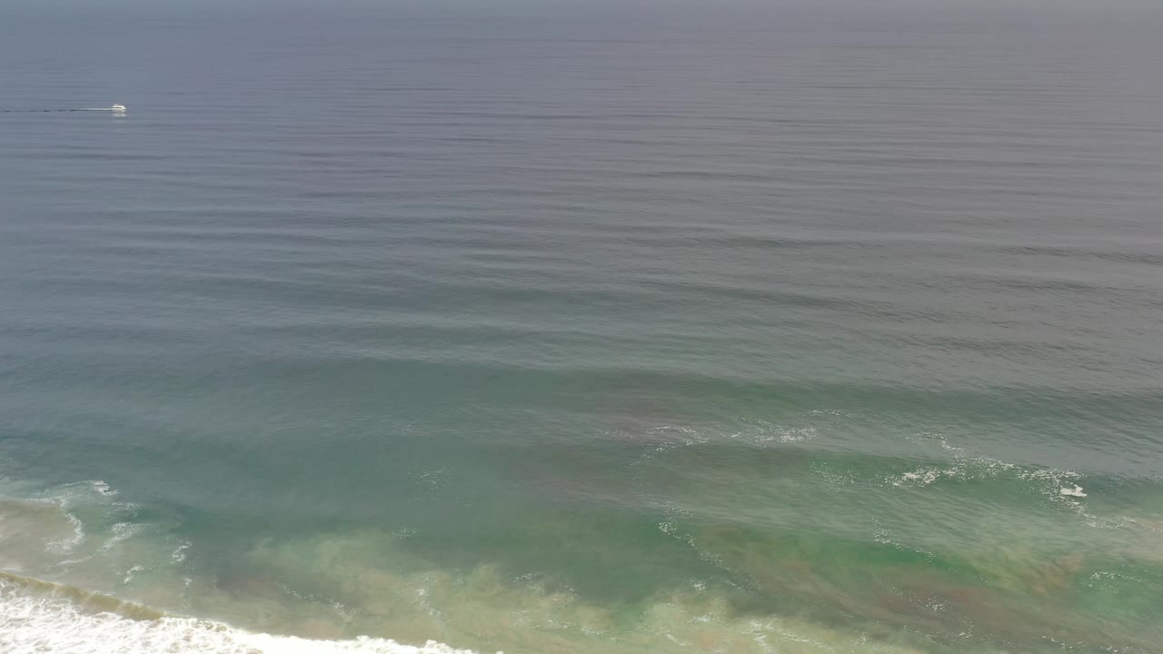 Green sea fills the Los Cabos frame with soft white wave lines at the lower edge, darker underwater patches through the midground, a hazy horizon, and no visible people.